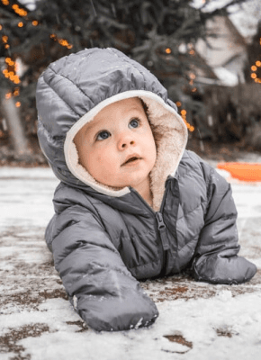 Baby in a gray winter coat crawling on snow-covered ground with holiday lights in the background
