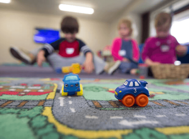 Children at kindergarten KIDI play on a colorful carpet with toy cars, which are in focus in the foreground while the kids sit blurred in the background.