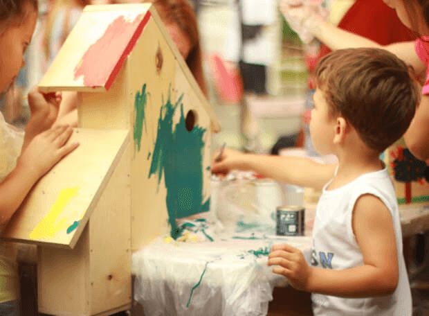 Children at kindergarten KIDI paint a large wooden birdhouse together, using bright colors like green, red, and yellow during a group art activity.